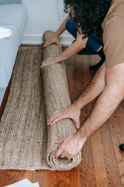 A person is actively rolling out a beige, textured area rug onto a polished wooden floor in a living room, with another individual assisting nearby. The room features a light-colored sofa with a white