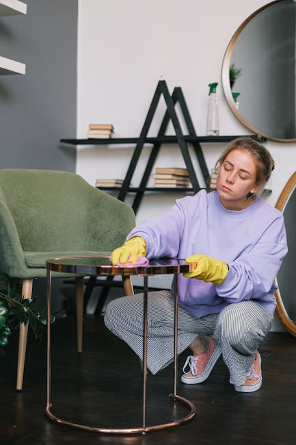 A person is actively rolling out a beige, textured area rug onto a polished wooden floor in a living room, with another individual assisting nearby. The room features a light-colored sofa with a white